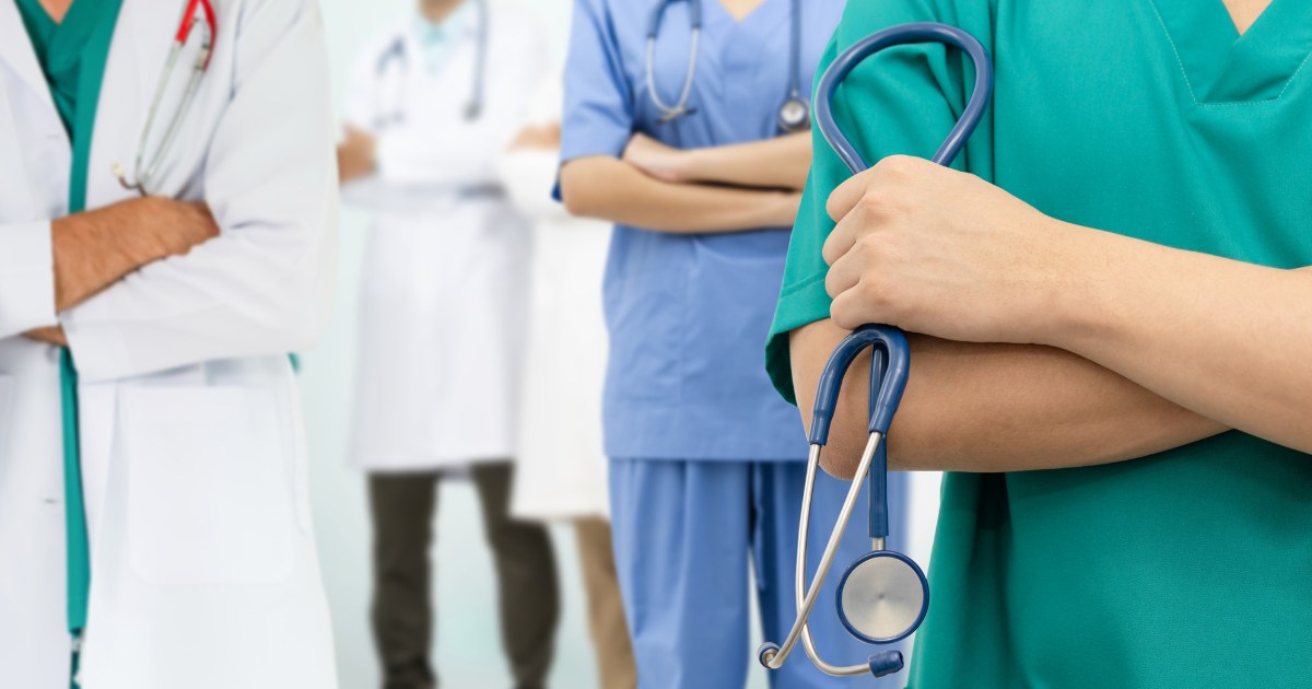 Nella foto, quattro operatori sanitari, indossando abiti da lavoro, sono in piedi con le braccia incrociate. In primo piano, una persona con uno stetoscopio appeso al collo indossa una divisa verde. Altri medici e infermieri con divise bianche e blu sfocate sono visibili sullo sfondo.
